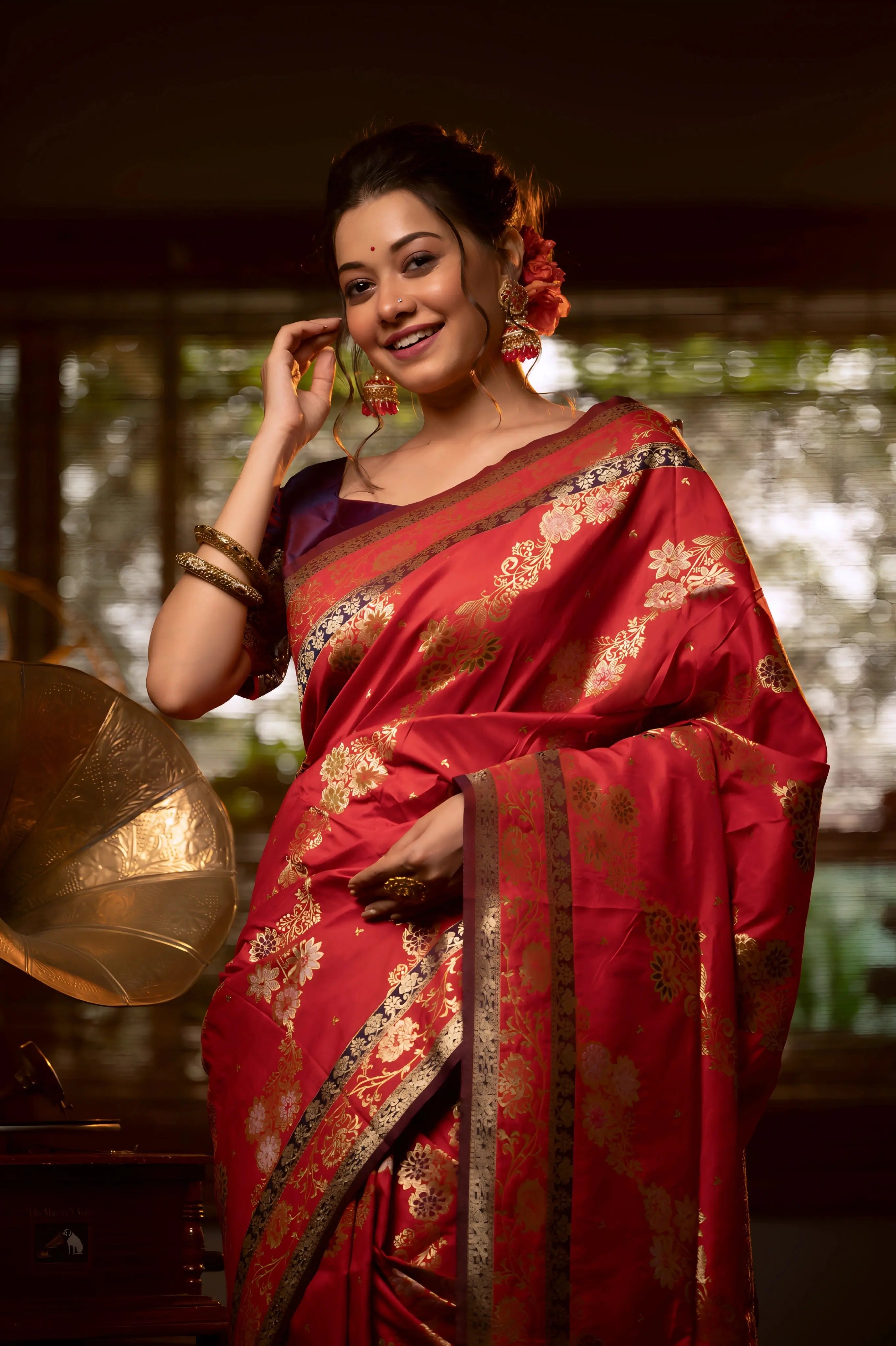 A woman models a luxurious Deep Red Soft Banarasi Silk Saree. The saree features a wide border and pallu with dense, intricate gold Zari weaving in traditional floral and paisley patterns. She wears a dark contrasting blouse and large gold jewelry, posing indoors on a black and white checkered floor.